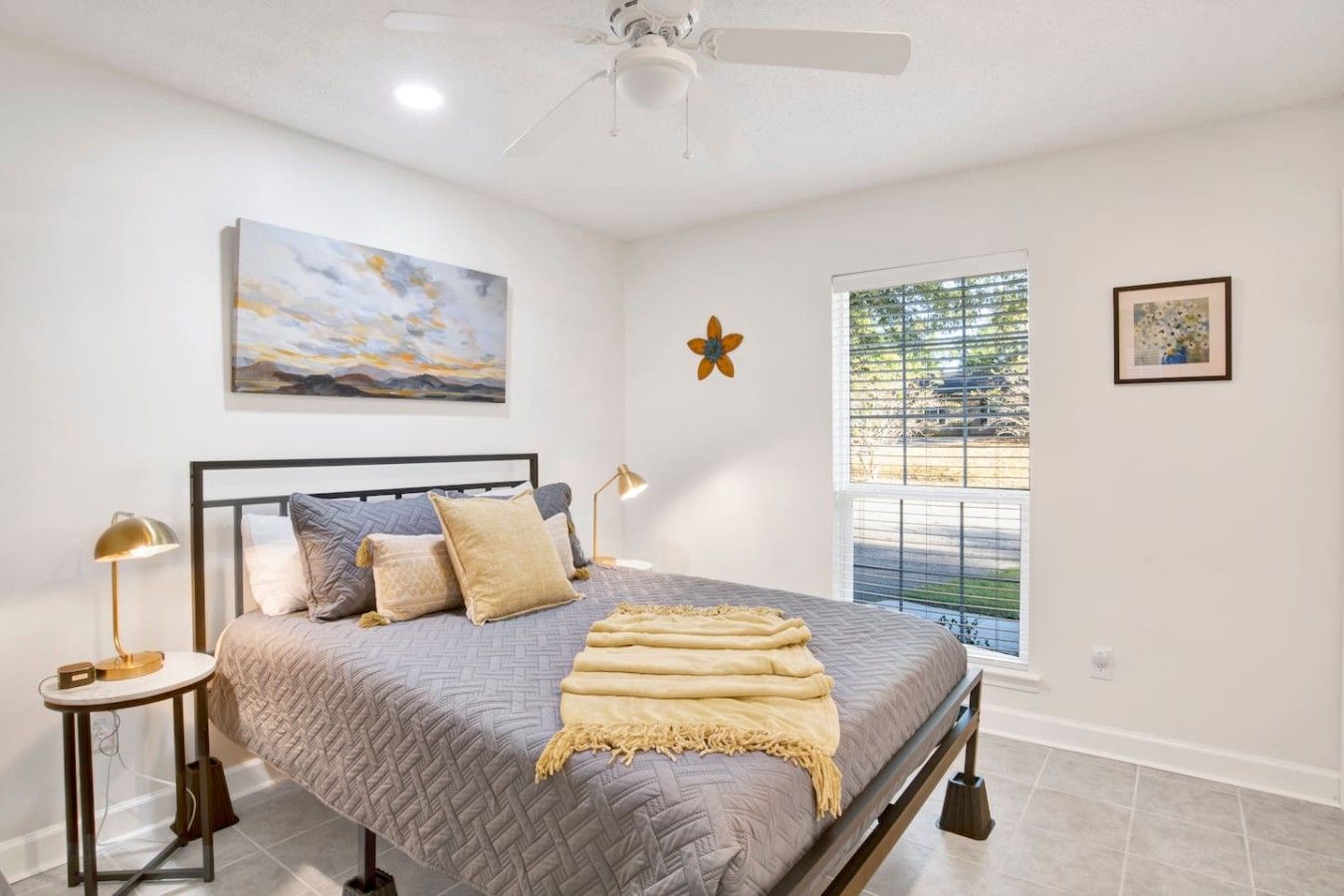 A bedroom with a bed, a nightstand, a lamp, a ceiling fan, a window with blinds, and artwork on the walls.
