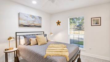 A bedroom with a bed, a nightstand, a lamp, a ceiling fan, a window with blinds, and artwork on the walls.
