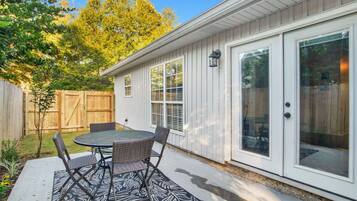 A patio with a round table and chairs, a wooden fence, and a house with white siding and large windows.