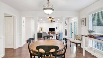 A spacious dining area with a large wooden table and chairs, a ceiling fan, and a view of the living room through an open doorway.