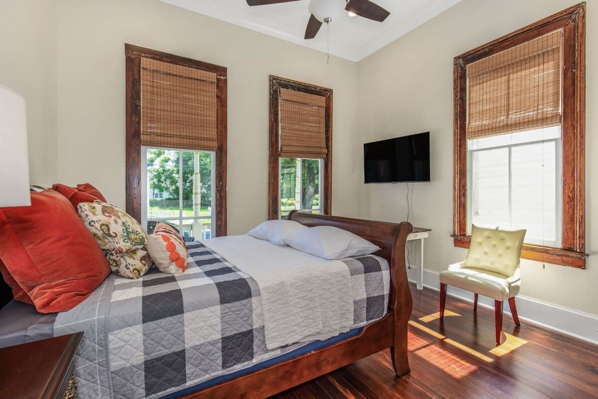 A bedroom with a bed, a chair, a television, and windows with blinds.