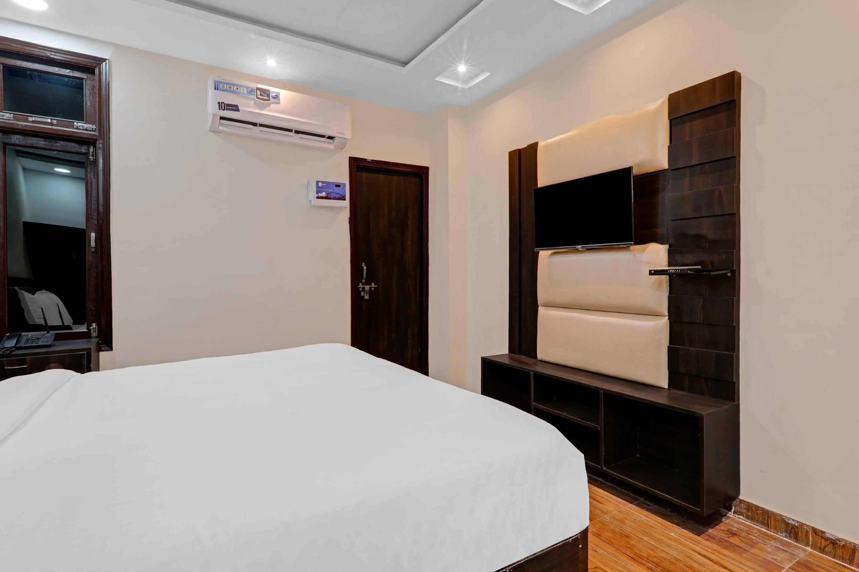 Une chambre d’hôtel comprenant un lit, un téléviseur à écran plat fixé sur un panneau en bois foncé, un climatiseur mural et une porte dotée d’une poignée.