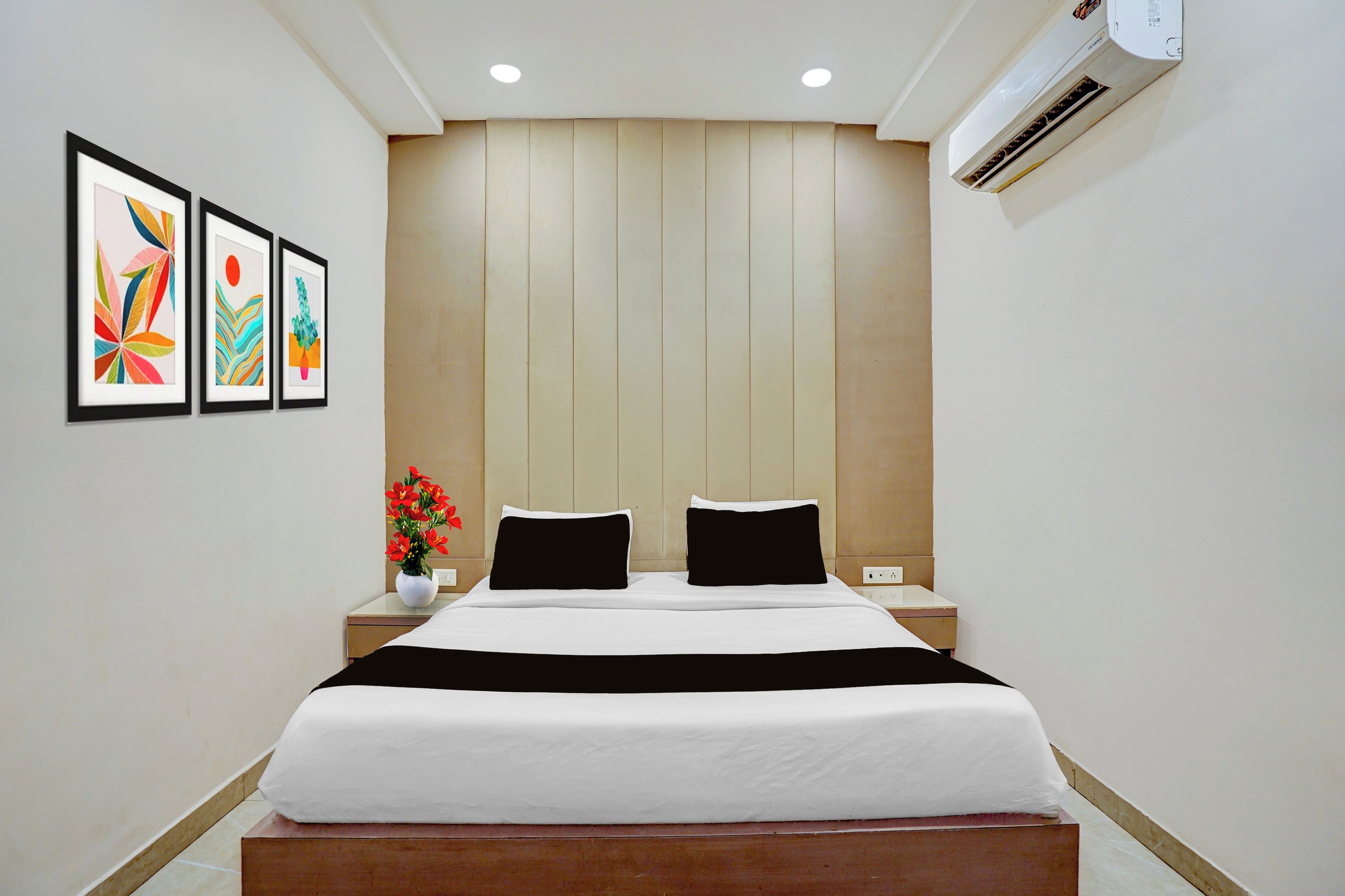 Una habitación de hotel con una cama, dos mesitas de noche, una mesita con un florero y tres cuadros en la pared.