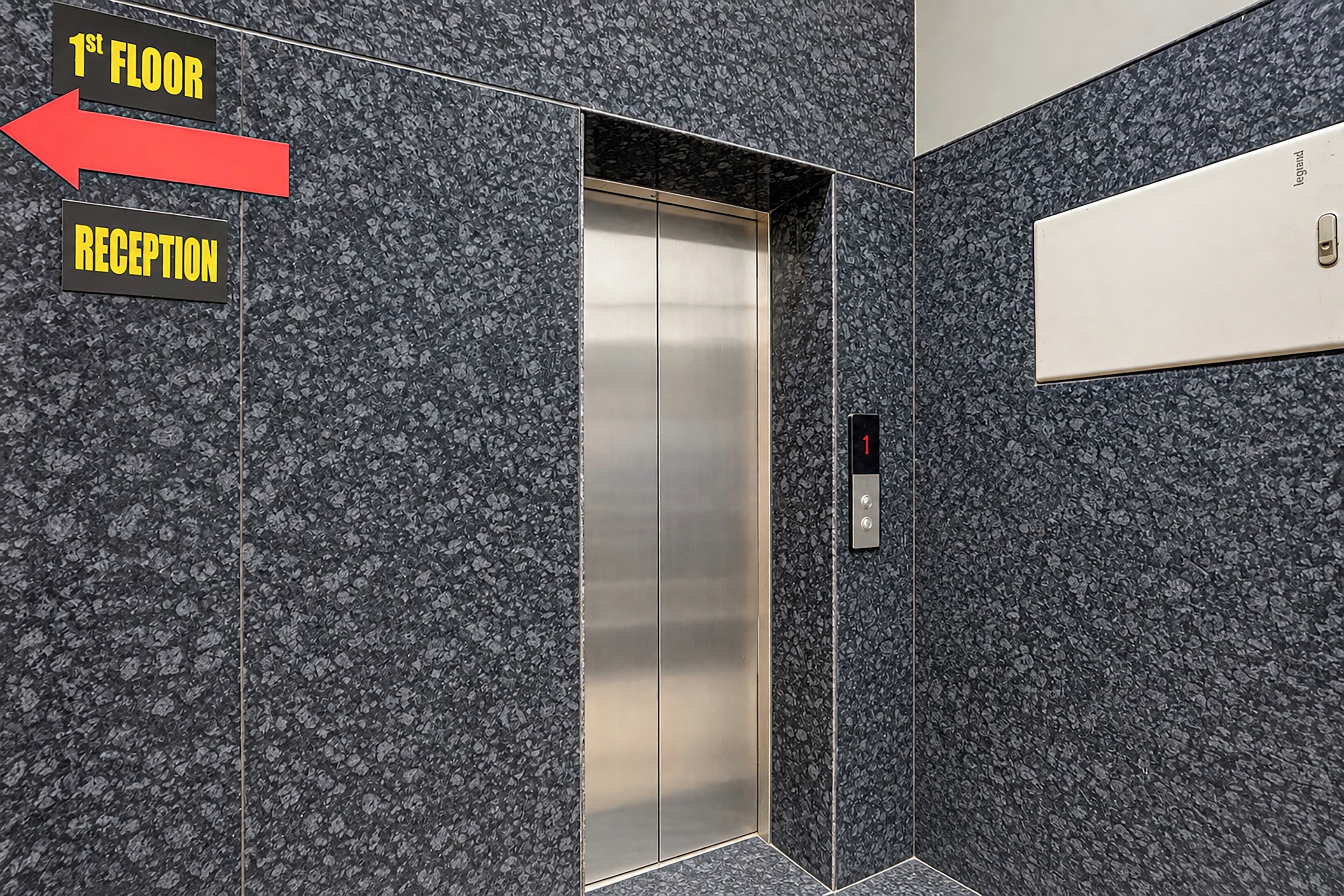 Un ascensor con interior metálico y un panel de control en la pared. Una señalización indica la ubicación de la recepción en la primera planta.
