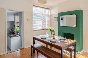 A dining area with a wooden table set for four, a bench, and a green accent wall with a mirror. - Swift Host | Albany House | Sleeps 4 | Parking | Garden | Good Location (Norwich)