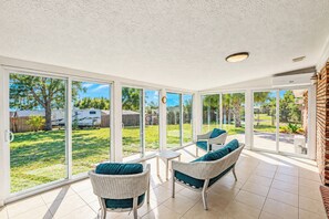 A sunlit patio with two wicker chairs and a small table, large sliding glass doors leading to a backyard with a lawn and a trailer. - Beautifully Decorated 4-bedroom house with Waterviews 7 minutes to Tyndall AFB (Panama City)