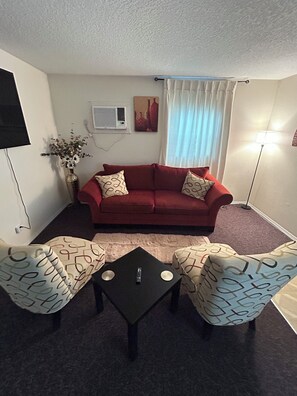 A living room with a red sofa, two patterned armchairs, a small black table, a floor lamp, a TV mounted on the wall, and a painting above the sofa. - The Haven - Cozy 1 bedroom apt near Busch Gardens and minutes from Tampa venues (Tampa)