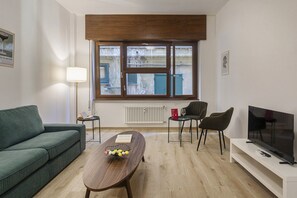 A modern living room with a green sofa, a wooden coffee table with a book and flowers, a TV on a stand, and a window with shutters. - XXV Aprile By Wonderful Italy - Menta (Genoa)