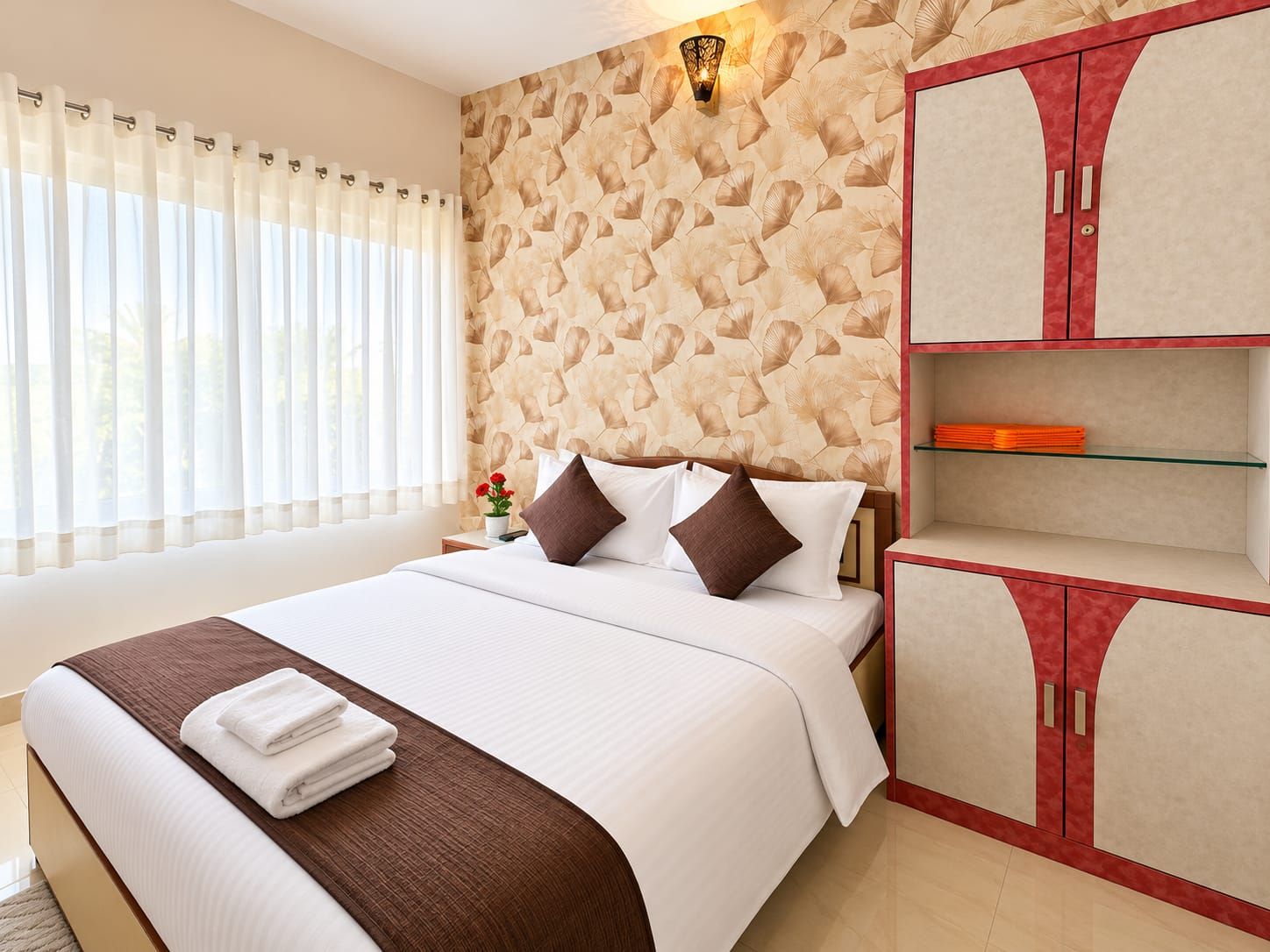Una habitación de hotel con cama, mesita de noche, pared con diseño de hojas, ventana con cortinas y armario con detalle rojo.