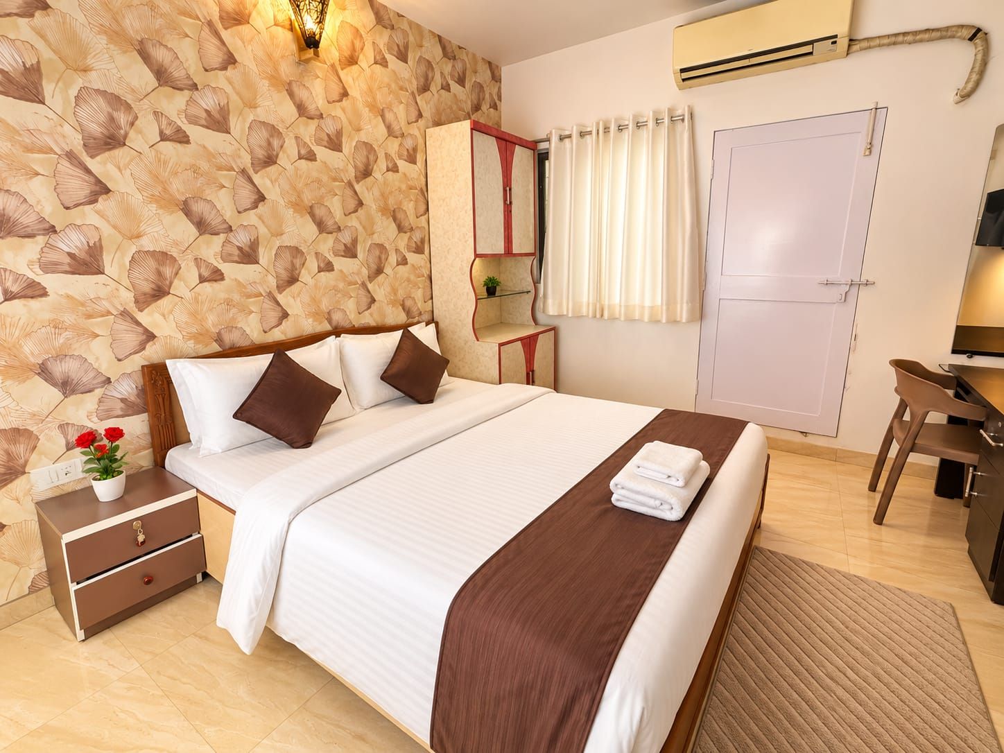 Quarto de hotel com cama, mesa de cabeceira, abajur, aparelho de ar condicionado e escrivaninha com cadeira.