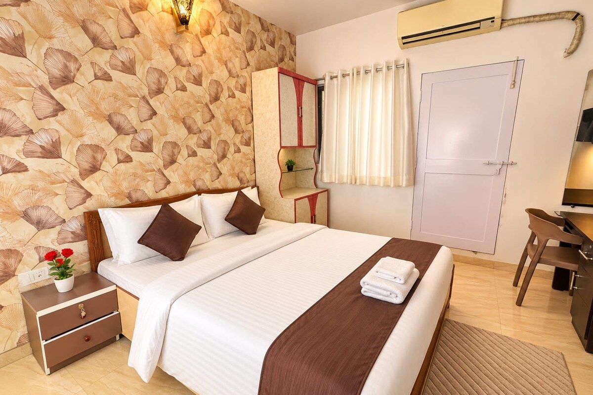 A hotel room with a bed, bedside table, lamp, air conditioning unit, and a desk with a chair.