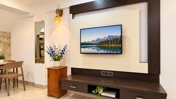 Una sala moderna con un televisor de pantalla plana montado en una pared de madera oscura, un mueble de madera pequeño, una planta en maceta y un comedor con sillas.