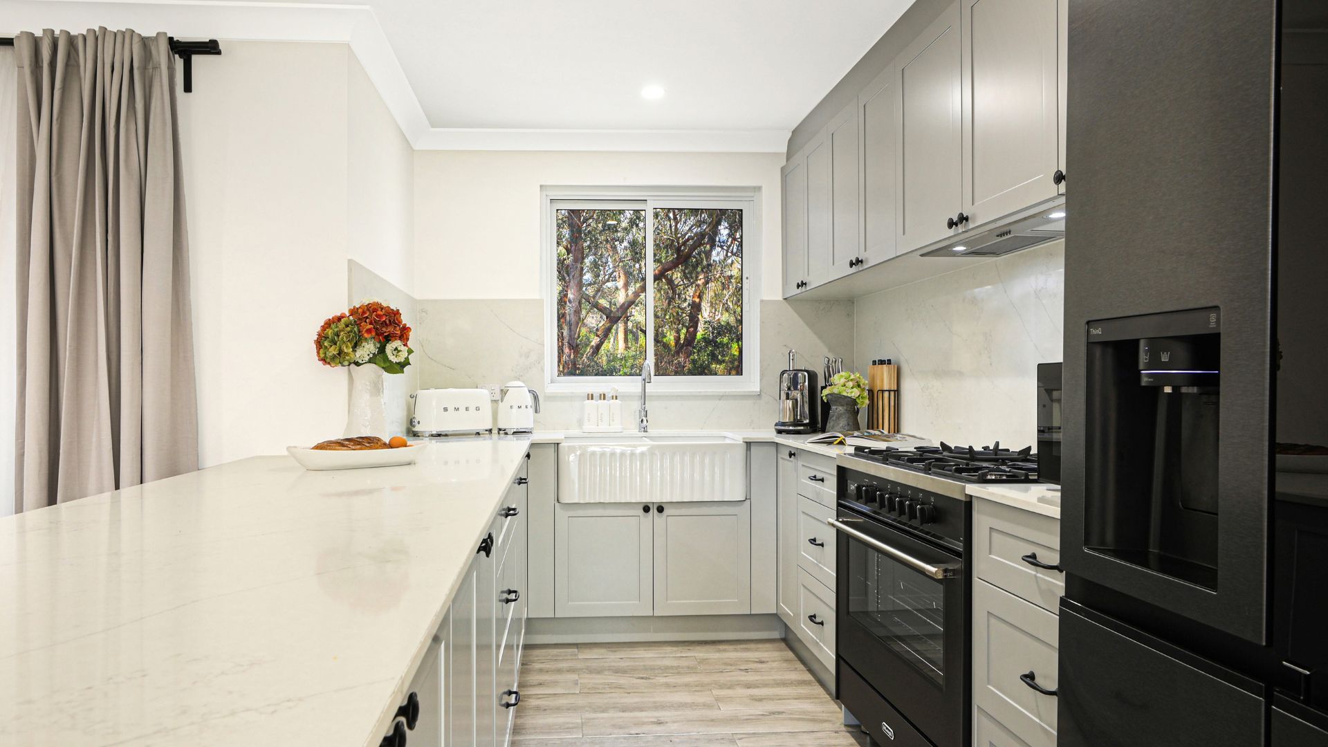 A modern kitchen with a white countertop, stainless steel appliances, and a window with a view of trees.
