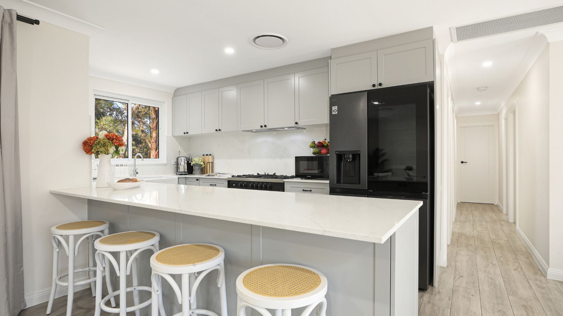 Una cocina moderna con armarios blancos, una isla grande con taburetes de bar, un refrigerador y una ventana con vistas a árboles.