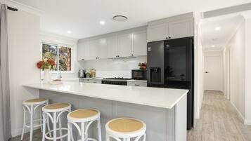 Una cocina moderna con armarios blancos, una isla grande con taburetes de bar, un refrigerador y una ventana con vistas a árboles.