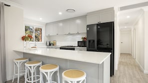 Une cuisine moderne avec des meubles blancs, un grand îlot central doté de tabourets, un réfrigérateur et une fenêtre donnant sur la verdure.