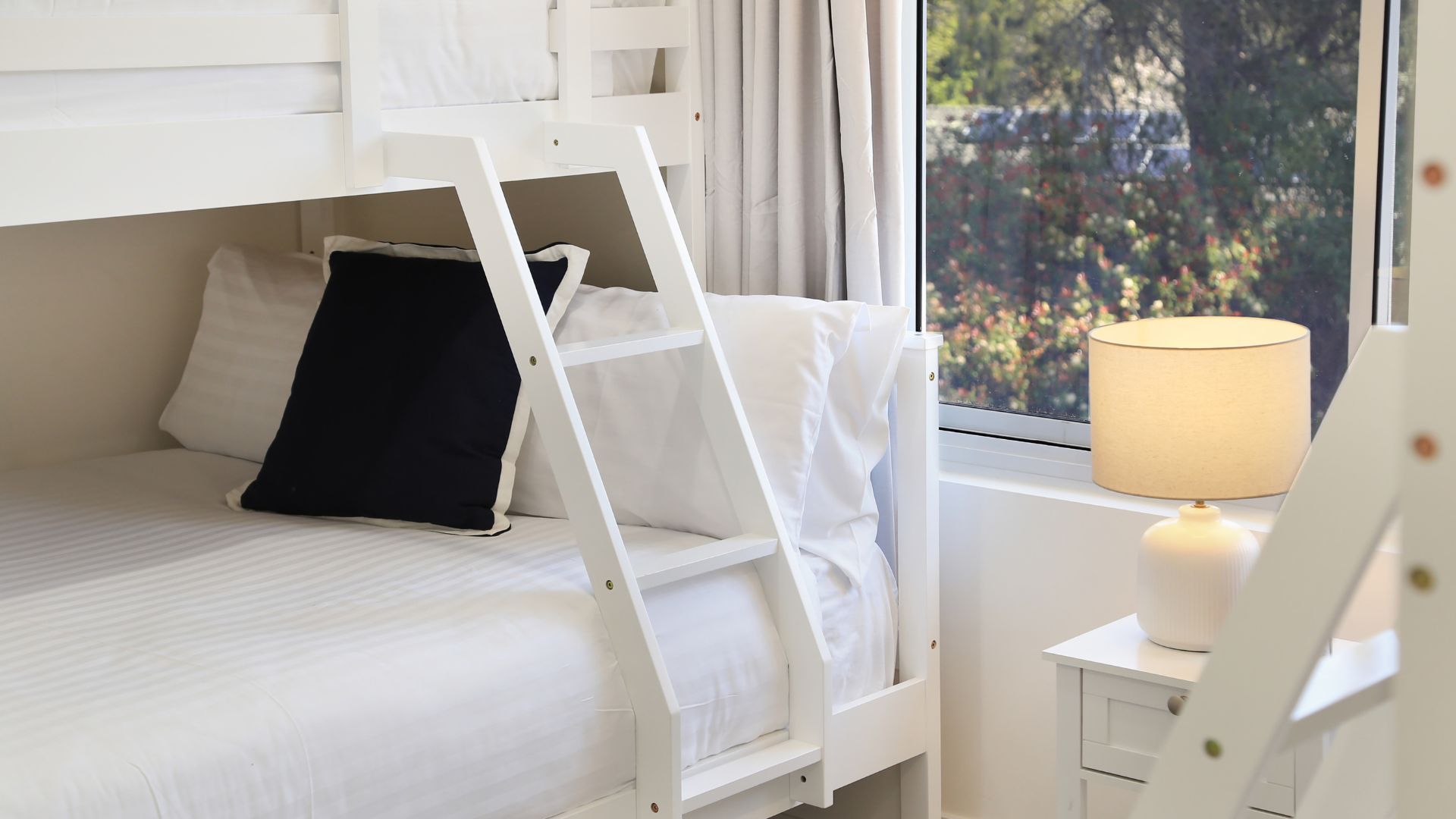 Una cama individual superpuesta con escalera, una mesita de noche con lámpara, y una ventana con vistas a la vegetación.