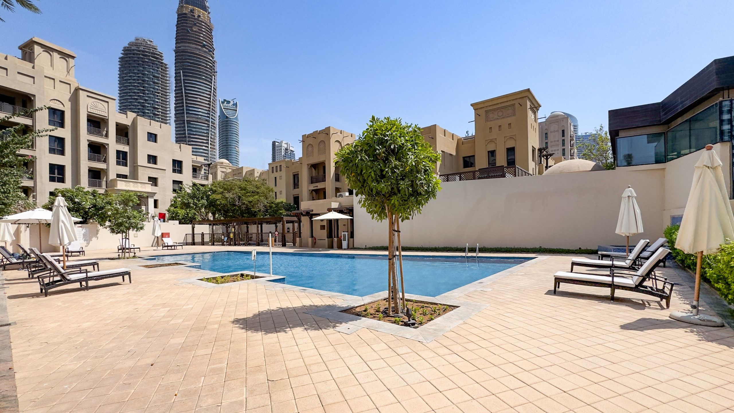 A rooftop swimming pool with lounge chairs and umbrellas, surrounded by buildings and a clear sky.