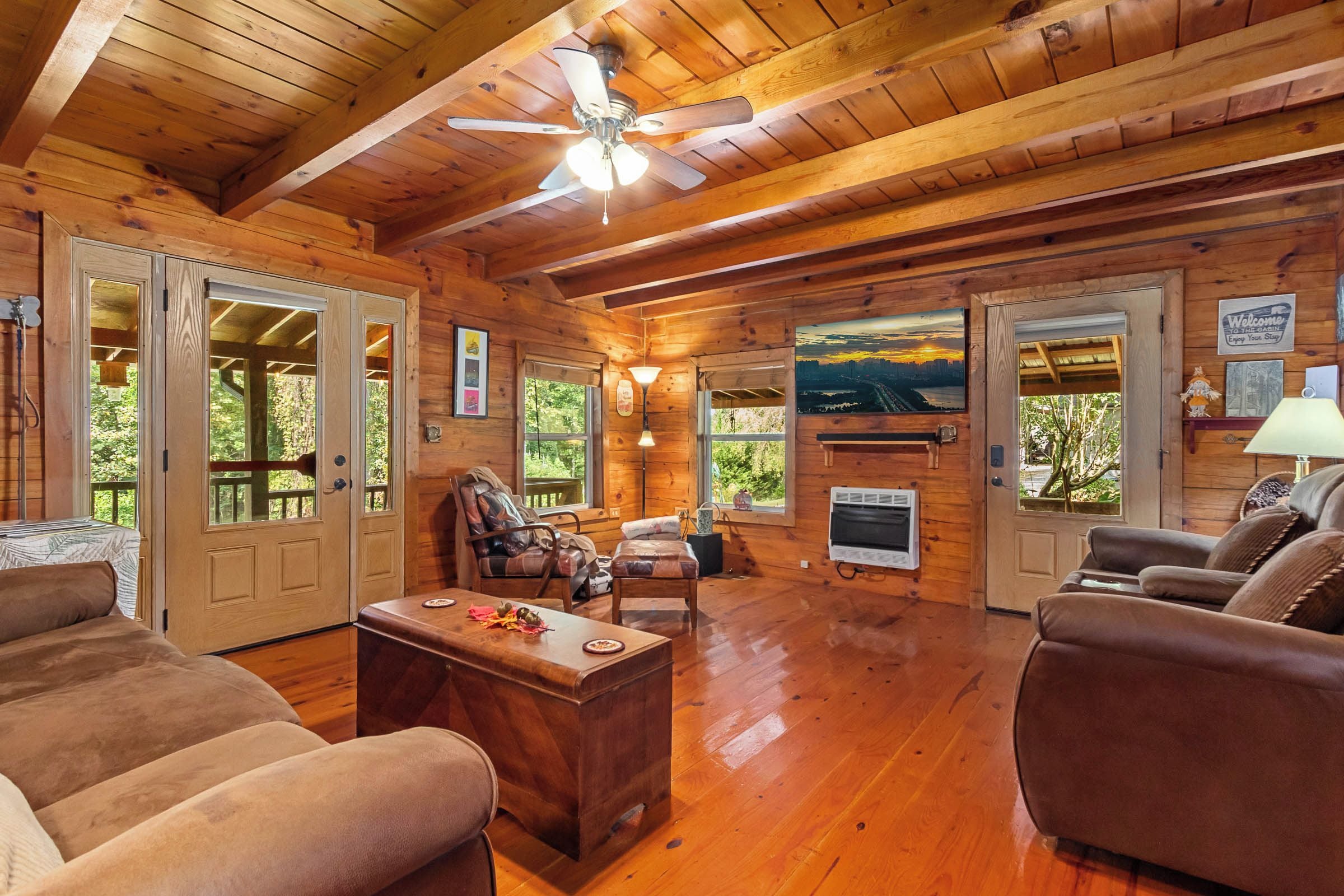 A cabin interior with wooden walls, a fireplace, a ceiling fan, and a view of the outdoors.