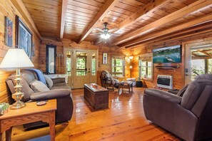 A cozy cabin interior with wooden floors, a ceiling fan, a fireplace, and a large window with a view of a river. - Cozy 3BR Mountain Cabin | Peaceful & Relaxing (Murphy)