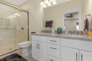 Un baño moderno con cabina de ducha de vidrio, inodoro blanco, tocador con encimera de mármol y ventilador de techo.