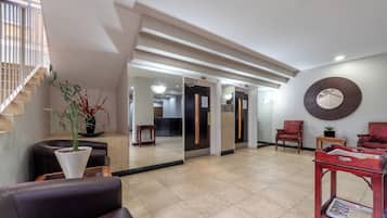 Um hall com escada, balcão de recepção, área de estar com cadeira e mesa, e um espelho grande na parede.