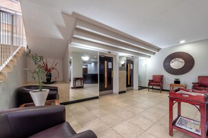 Un vestíbulo con una escalera, una recepción, un área de descanso con una silla y una mesa, y un espejo grande en la pared.
