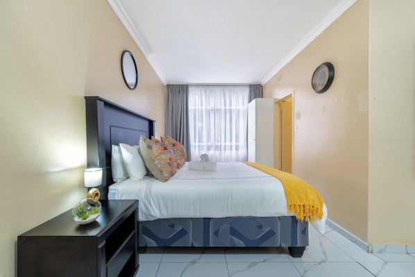 A hotel room with a bed, a nightstand, a lamp, a window with curtains, and a door.