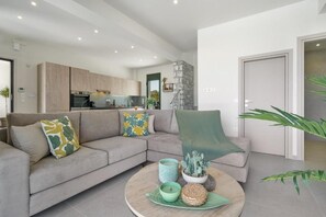 A modern living room with a grey sofa, a round coffee table, and a kitchen in the background. - Villa Meraki Living in Nature with Heated Pool (Stalos)