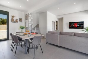 A modern living room with a grey sofa, a dining table with chairs, a TV mounted on the wall, and a staircase leading to another level. - Villa Meraki Living in Nature with Heated Pool (Stalos)