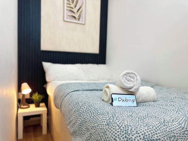 A neatly made bed with rolled towels and a sign, a bedside lamp, and a small plant on a nightstand. - Quiet Partition Room in Tecom Near Metro (Dubai)