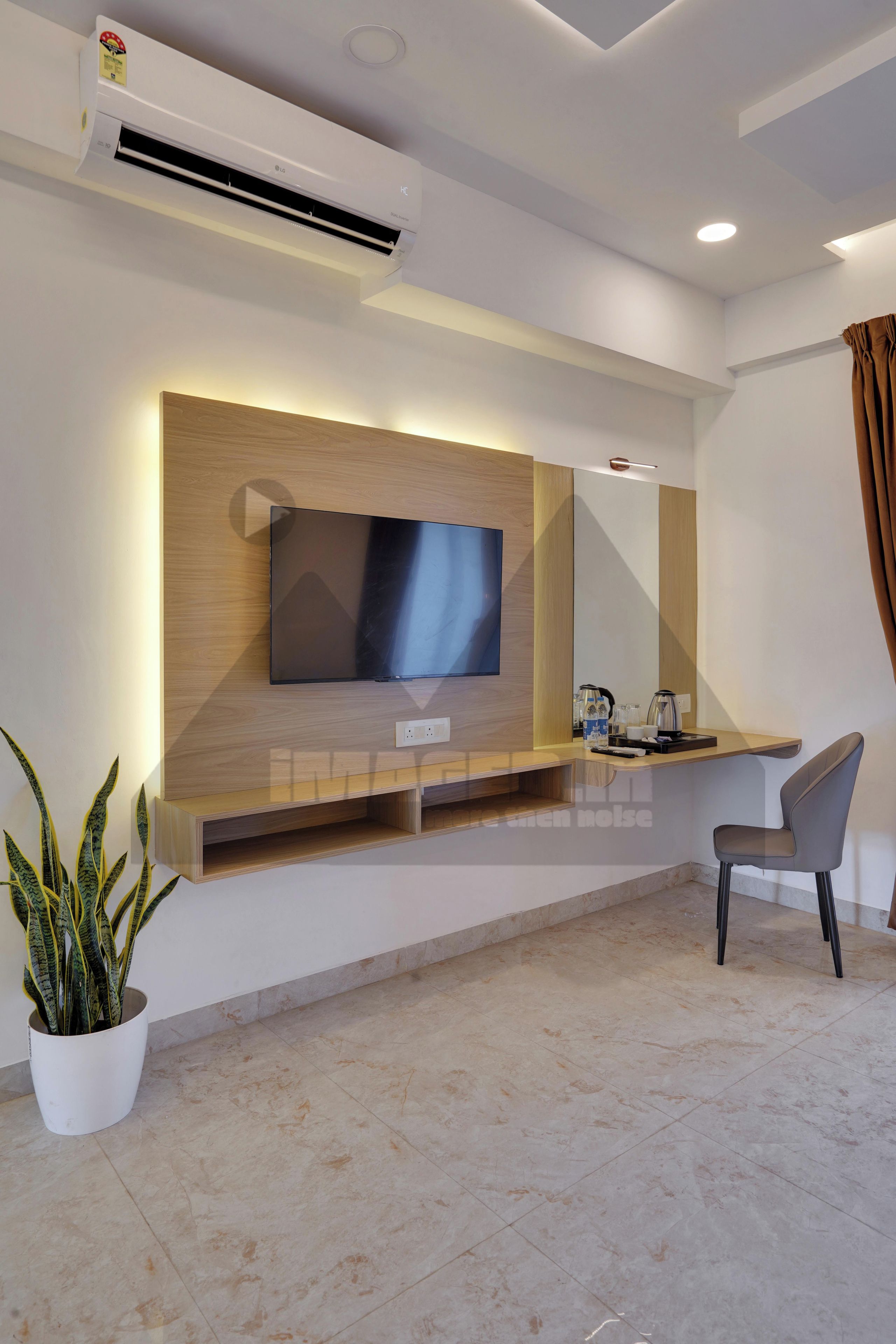 Een moderne hotelkamer met een flatscreen televisie bevestigd aan een houten paneel, een klein bureau met een stoel, een potplant en een betegelde vloer.