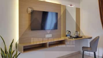 Een moderne hotelkamer met een flatscreen televisie bevestigd aan een houten paneel, een klein bureau met een stoel, een potplant en een betegelde vloer.