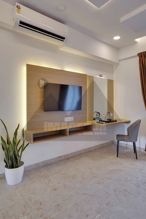 Habitación de hotel moderna con televisión de pantalla plana montada en un panel de madera, un escritorio pequeño con una silla, una planta en maceta y suelo de baldosas.