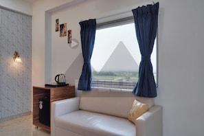 Habitación con sofá blanco, mesa de centro, hervidor y vista al campo a través de una ventana con cortinas azules.