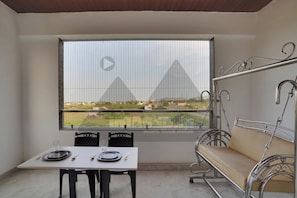 A room with a dining table set for two, a cushioned bench, and a large window offering a view of a landscape with buildings. - Hotel Regal Heaven (Sasvad)