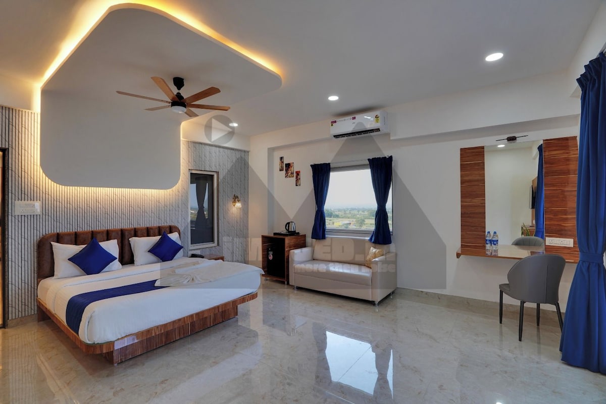 A modern hotel room with a large bed, a seating area, a desk, and a view of the outdoors.