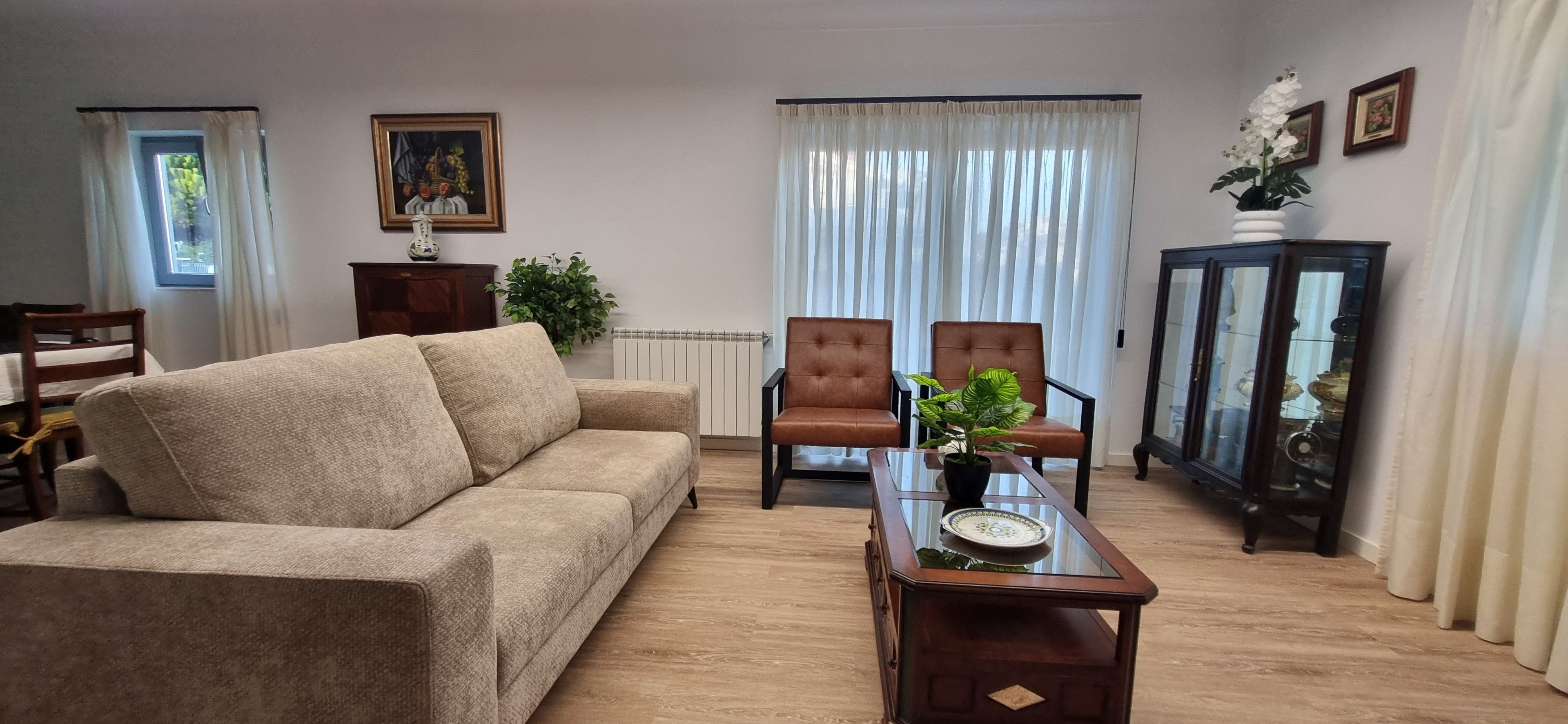 A living room with a beige sofa, two brown armchairs, a glass coffee table, and a cabinet with glass doors.