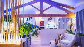Ein modernes Wohnzimmer mit hoher Decke, Holzbalken, einer großen Pflanze, einem weißen Sofa, einem kleinen Tisch mit Getränken und einem Fernseher.