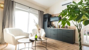 Ein modernes Wohnzimmer mit einer Couch, einem Couchtisch und einem Essbereich. Es gibt eine Küche mit dunklen Schränken und einem an der Wand montierten Fernseher. Eine große Pflanze ist in der Ecke zu sehen.