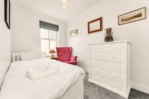 Een slaapkamer met een bed, een dressoir, een stoel, een raam met jaloezieën en aan de muur hangende foto's.