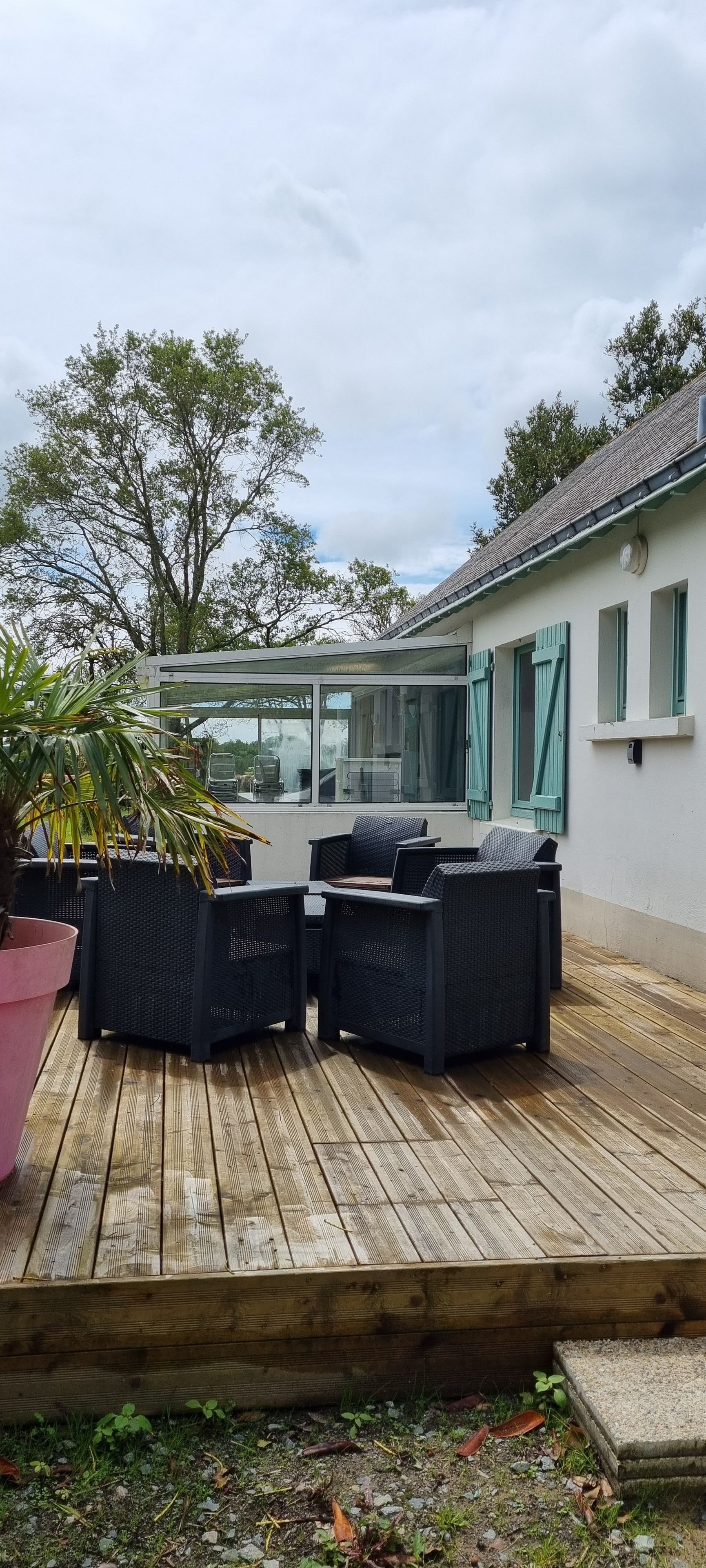 Una terraza de madera con mobiliario exterior, un cerramiento de vidrio y un edificio con ventanas.