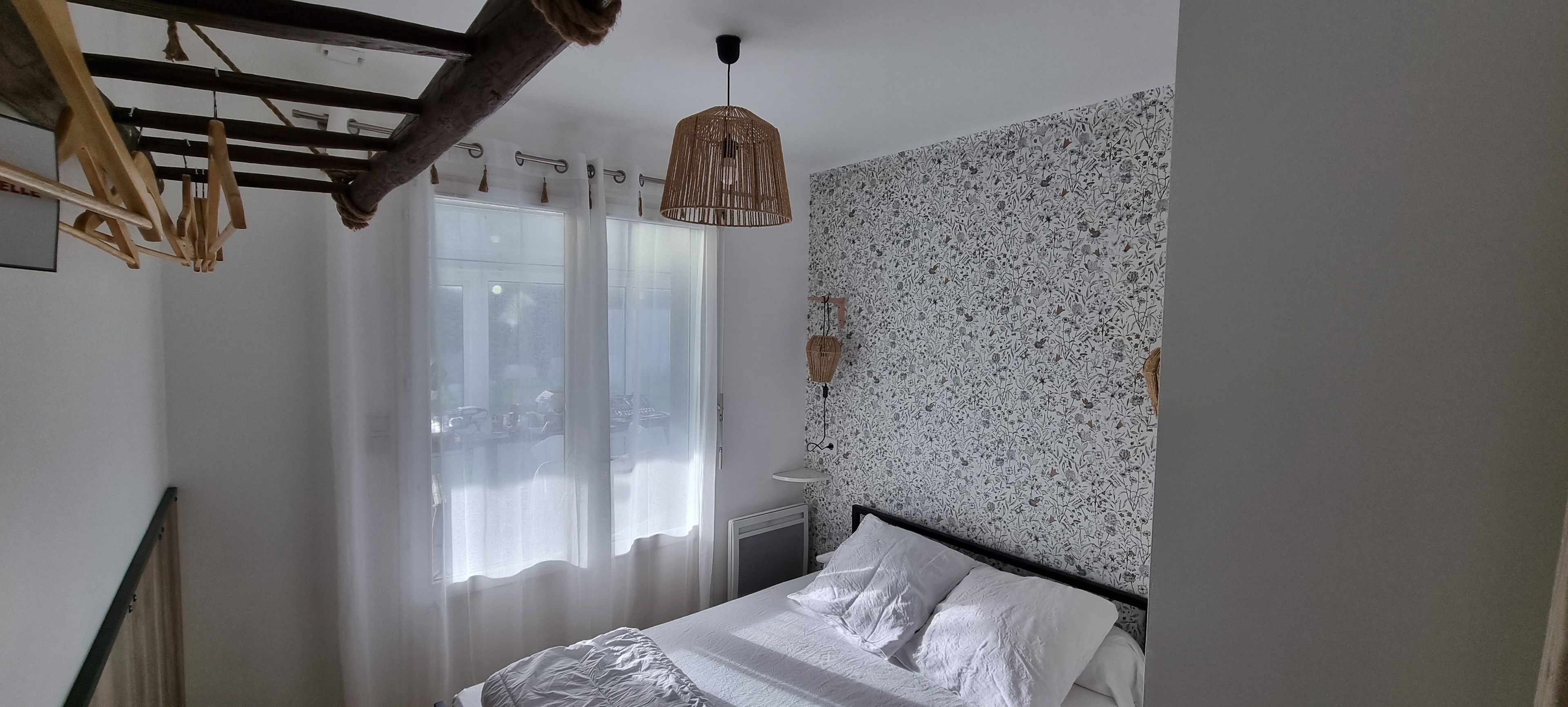 A bedroom with a bed, a window with sheer curtains, a wall with a floral pattern, and a hanging light fixture.
