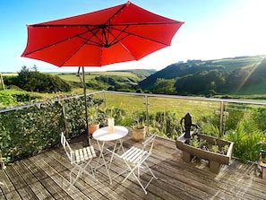 Eine Holzterrasse mit einem roten Sonnenschirm, weißen Stühlen und einem Tisch mit Blick auf ein Tal.