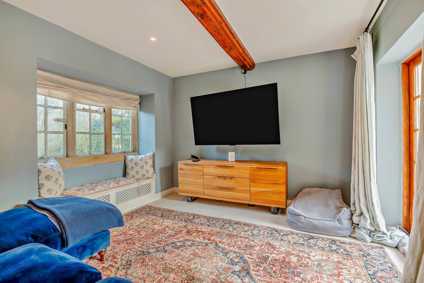 A bedroom with a large bed, a wooden dresser, a flat-screen TV, a window with curtains, and a patterned rug.