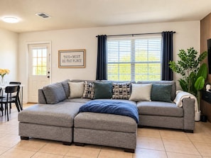 A living room with a sectional sofa, a dining table with chairs, a large window with curtains, and a framed quote on the wall. - 3BR Near Morgan Wonderland With Arcade (San Antonio)