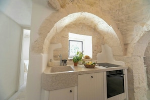 A kitchen with a stone archway, a window, a sink, a coffee maker, a potted plant, and a fruit basket. - Trulli complex in the Valle d'Itria with pool (Martina Franca)