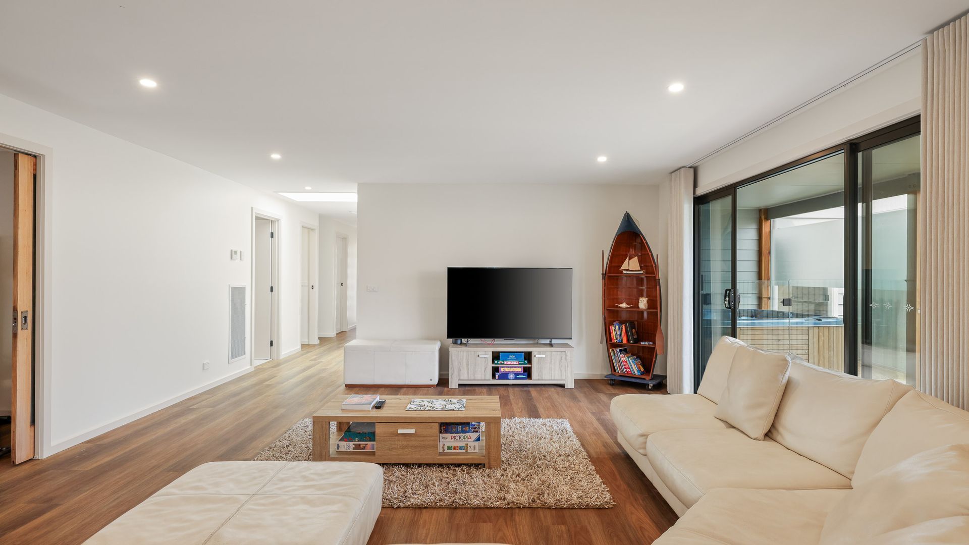 A modern living room with a flat-screen TV, a wooden coffee table, a beige sofa, and a large window offering a view of a balcony.