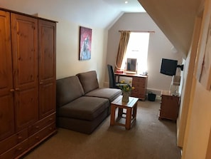 A cozy living room with a brown sofa, a small wooden table with a vase, a wooden cabinet, a television, and a window with curtains.