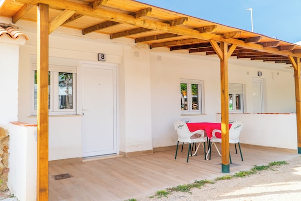 A covered outdoor seating area with a table and chairs, wooden beams overhead, and a door with a number.